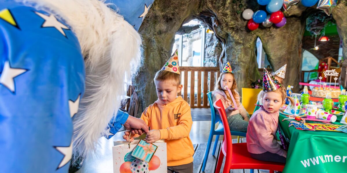 Eine Person in einem Zaubererkostüm überreicht einem Kind auf einer Geburtstagsparty eine Geschenktüte. Andere Kinder mit Partyhüten sitzen an einem geschmückten Tisch, über dem Luftballons schweben.