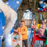Eine Person in einem Zaubererkostüm überreicht einem Kind auf einer Geburtstagsparty eine Geschenktüte. Andere Kinder mit Partyhüten sitzen an einem geschmückten Tisch, über dem Luftballons schweben.