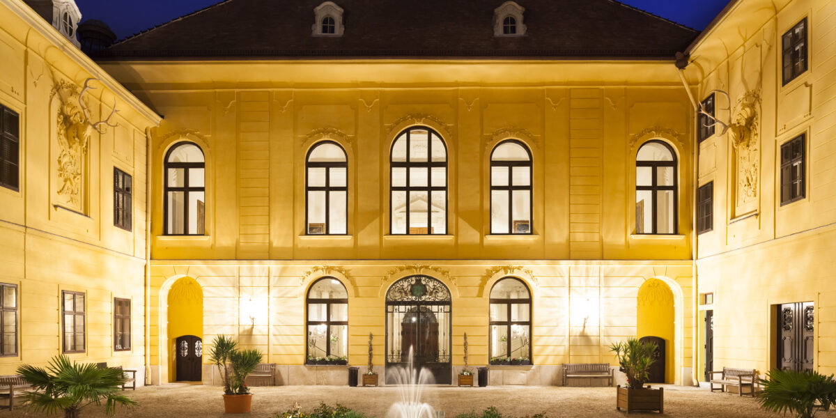 Der beleuchtete Innenhof eines gelben historischen Gebäudes bei Nacht, mit symmetrischen Fenstern und einem zentralen Springbrunnen, der von Pflanzen umgeben ist.