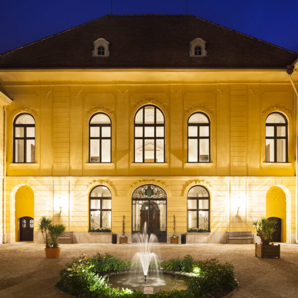 Der beleuchtete Innenhof eines gelben historischen Gebäudes bei Nacht, mit symmetrischen Fenstern und einem zentralen Springbrunnen, der von Pflanzen umgeben ist.