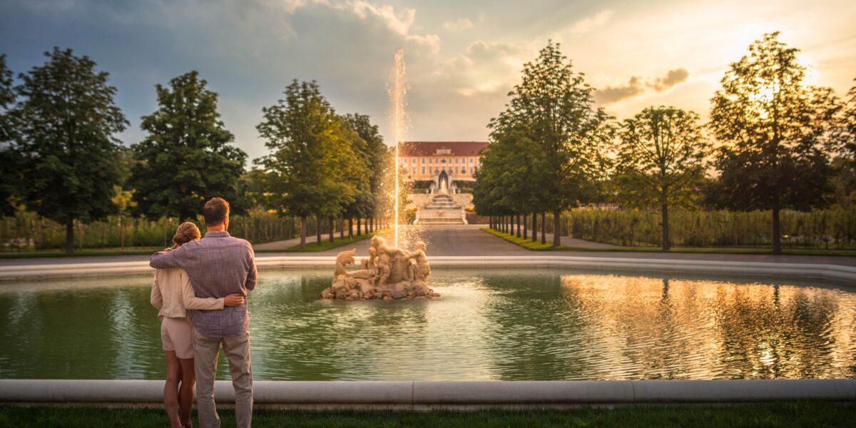 Ein Paar steht bei Sonnenuntergang neben einem Springbrunnen in einem parkähnlichen Garten und blickt auf ein großes historisches Gebäude in der Ferne.