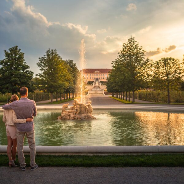 Ein Paar steht bei Sonnenuntergang neben einem Springbrunnen in einem parkähnlichen Garten und blickt auf ein großes historisches Gebäude in der Ferne.