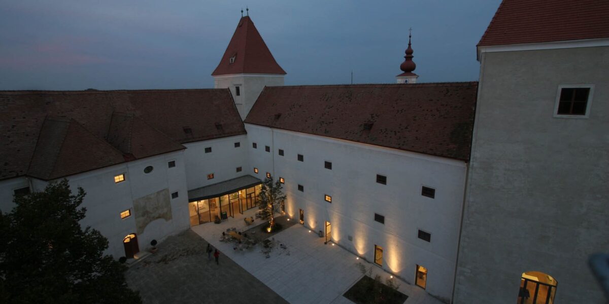 Ein großes historisches Gebäude mit roten Dächern und einem Turm, das in der Abenddämmerung in warmes Licht getaucht ist, umgibt einen Innenhof, auf dem sich einige Menschen aufhalten, sowie einen zentralen Gartenbereich.