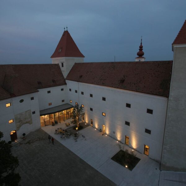 Ein großes historisches Gebäude mit roten Dächern und einem Turm, das in der Abenddämmerung in warmes Licht getaucht ist, umgibt einen Innenhof, auf dem sich einige Menschen aufhalten, sowie einen zentralen Gartenbereich.