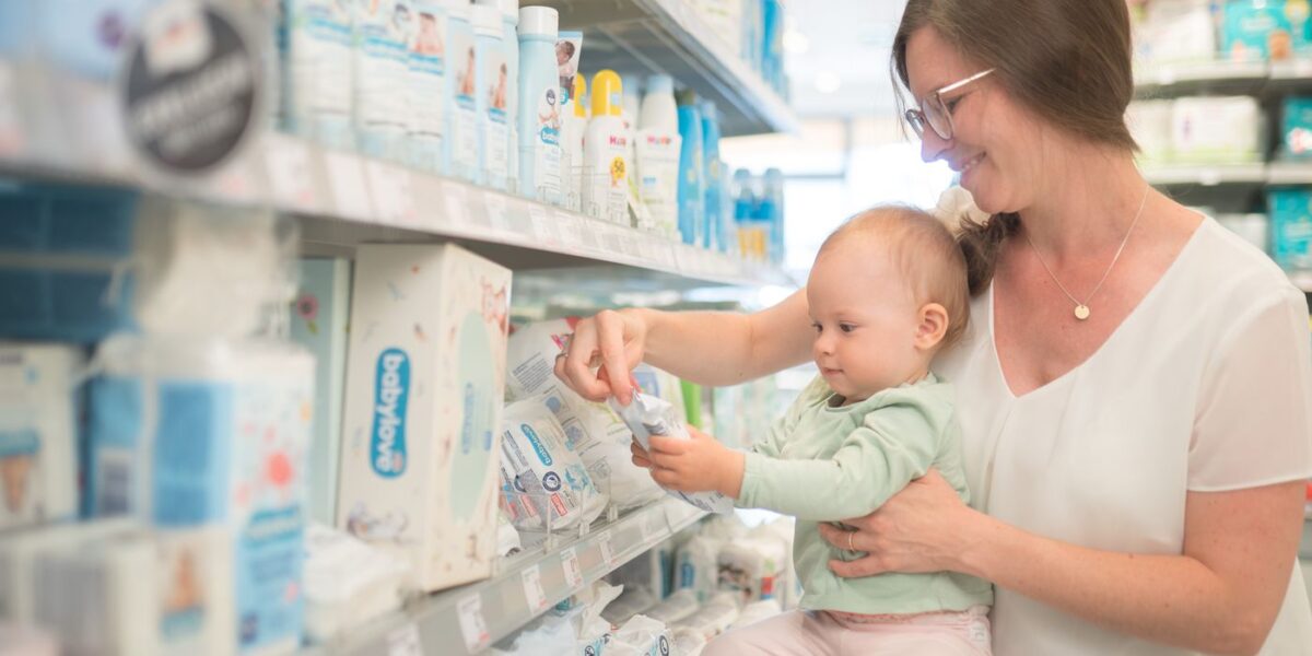 Eine Frau, die ein Baby im Arm hält, schaut sich in einem Ladenregal Babyartikel an und wählt einen Artikel aus dem Regal aus.