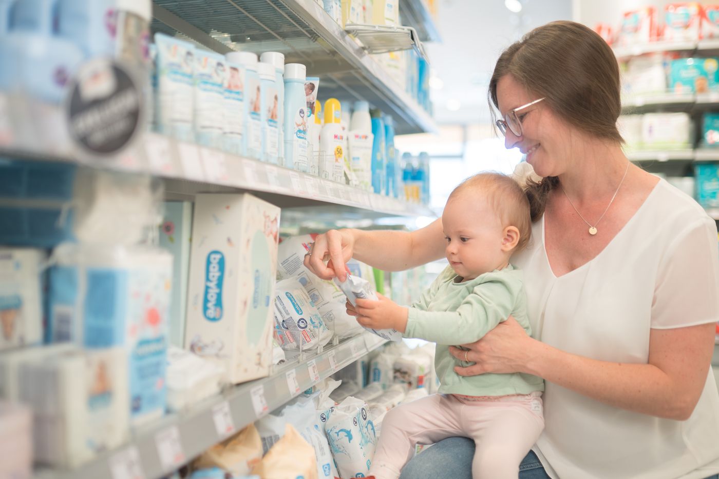 Eine Frau, die ein Baby im Arm hält, schaut sich in einem Ladenregal Babyartikel an und wählt einen Artikel aus dem Regal aus.