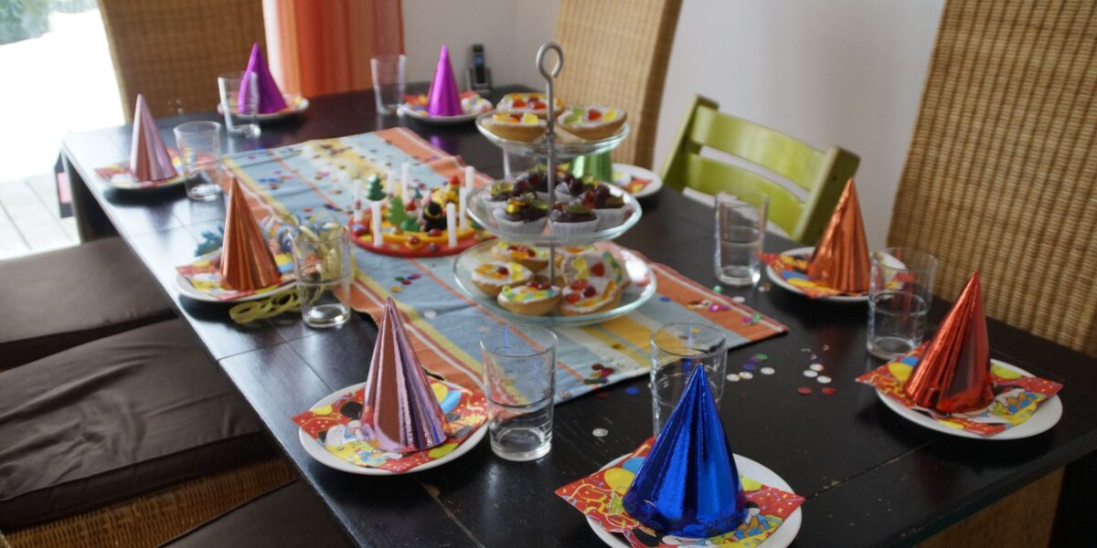 Ein für eine Party gedeckter Esstisch mit Partyhüten auf den Tellern, Gläsern, bunten Servietten und Tabletts mit verschiedenen Snacks in der Mitte.