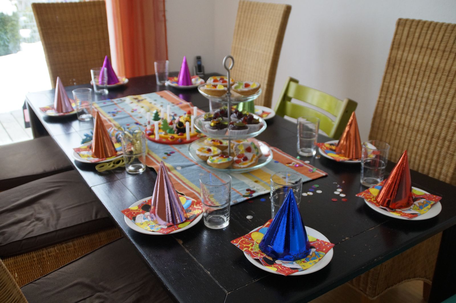 Ein für eine Party gedeckter Esstisch mit Partyhüten auf den Tellern, Gläsern, bunten Servietten und Tabletts mit verschiedenen Snacks in der Mitte.