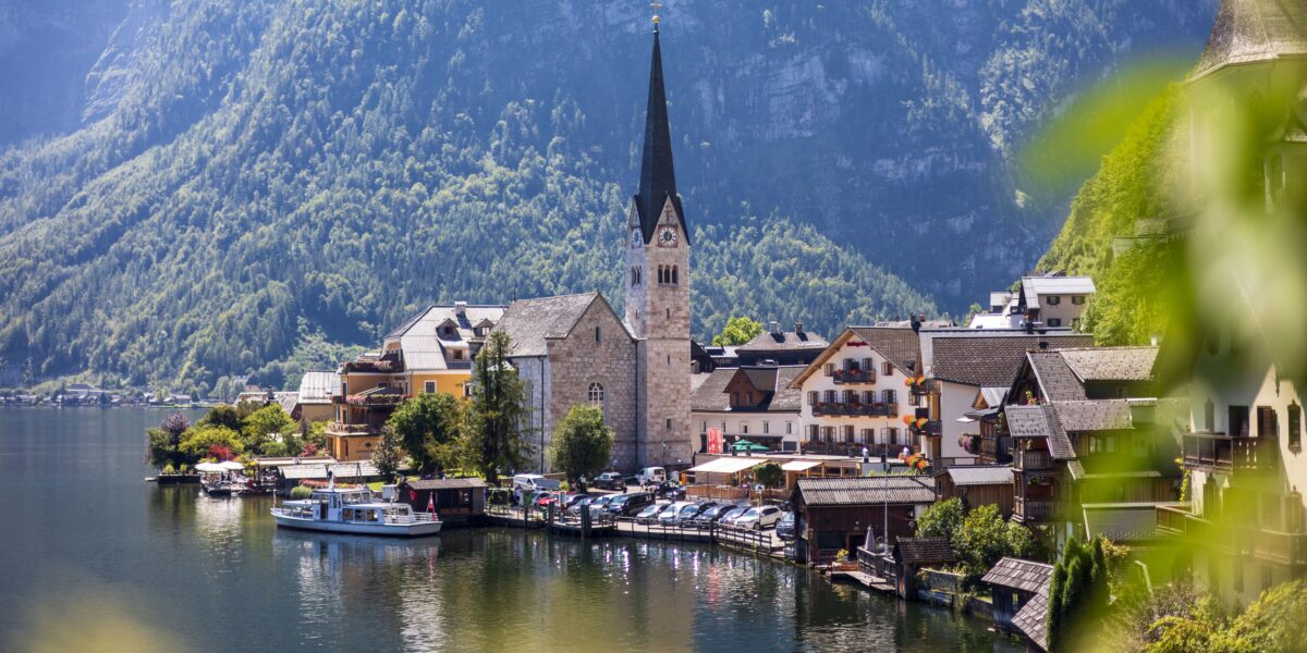Ein Dorf am See mit einem Kirchturm, traditionellen Gebäuden, am Ufer liegenden Booten und üppig grünen Bergen im Hintergrund an einem sonnigen Tag.