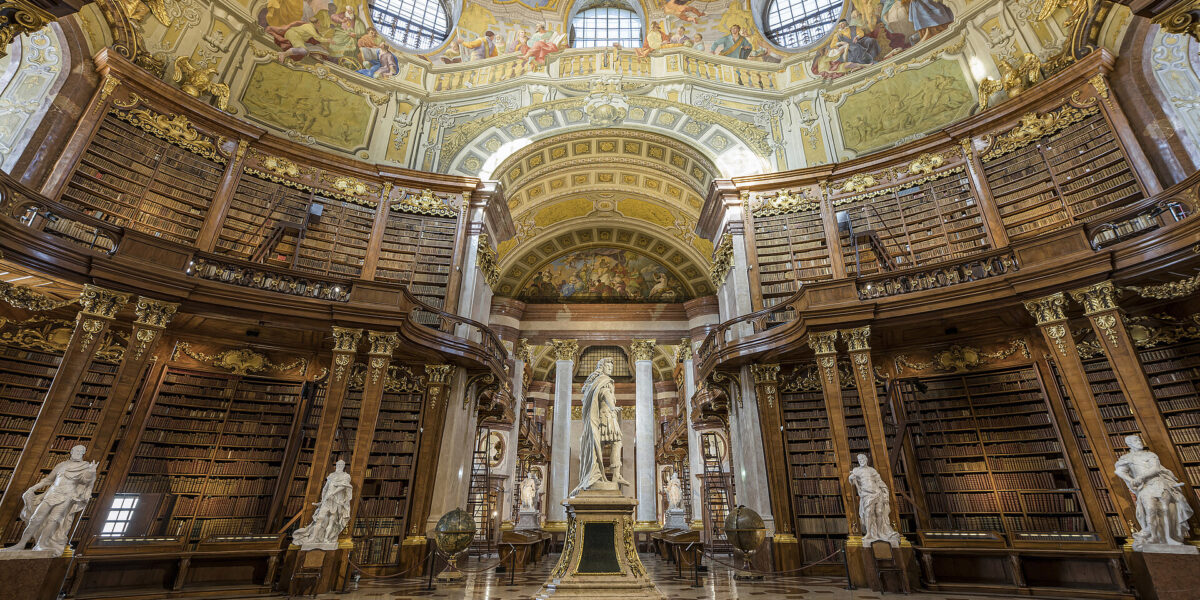 Ein prunkvolles, historisches Bibliotheksinterieur mit hohen Holzregalen, Marmorstatuen und einer gewölbten, mit Fresken verzierten Decke, die durch große Bogenfenster und aufwendige Kunstwerke besticht.