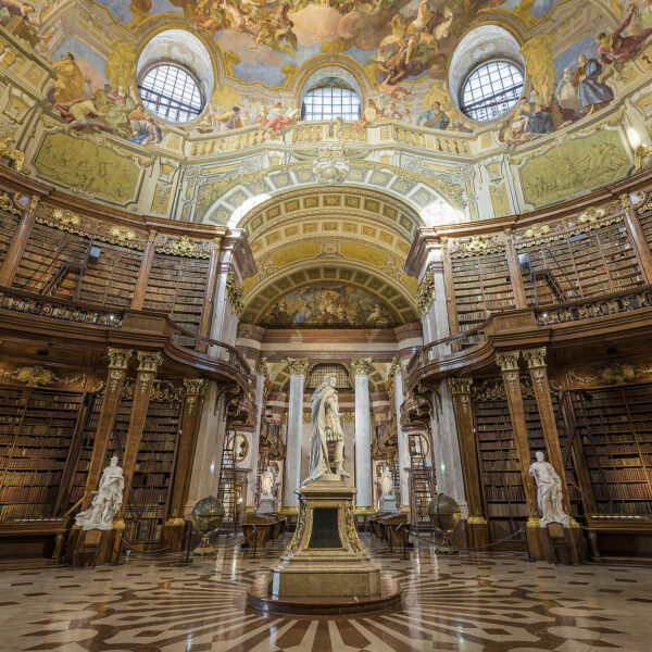 Ein prunkvolles, historisches Bibliotheksinterieur mit hohen Holzregalen, Marmorstatuen und einer gewölbten, mit Fresken verzierten Decke, die durch große Bogenfenster und aufwendige Kunstwerke besticht.