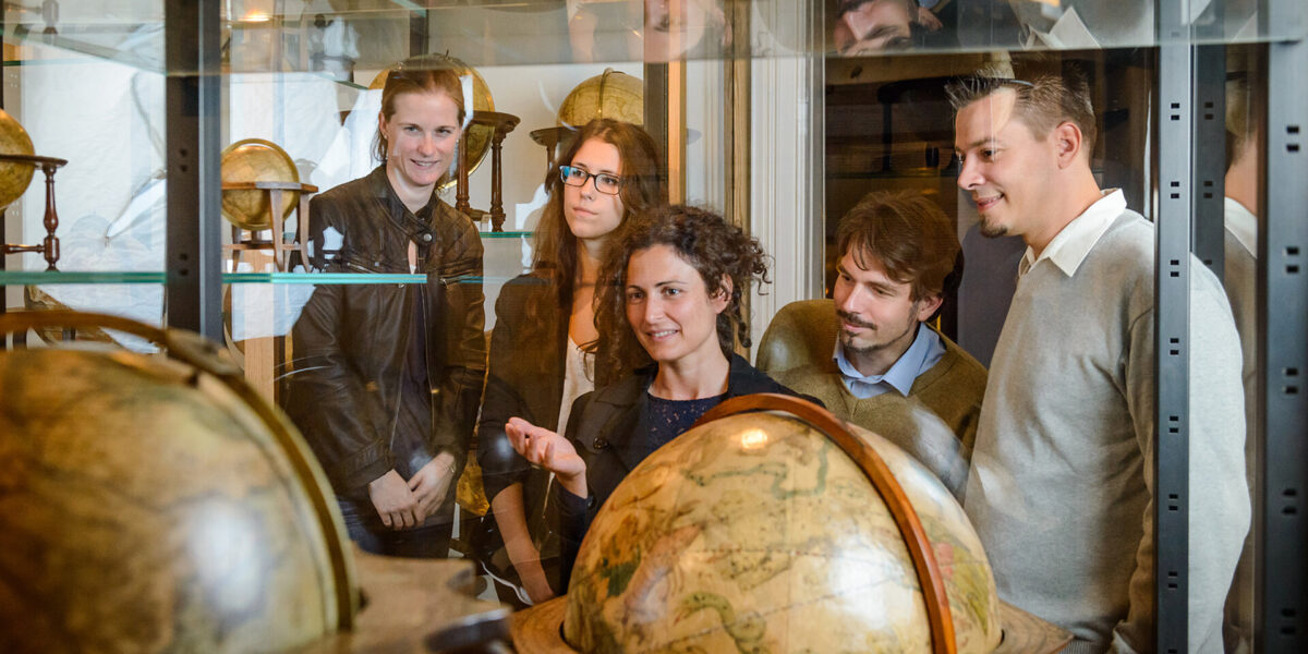 Fünf Personen stehen in einem Museum, betrachten antike Globen, die in Vitrinen ausgestellt sind, und unterhalten sich darüber.
