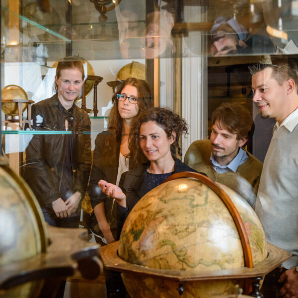 Fünf Personen stehen in einem Museum, betrachten antike Globen, die in Vitrinen ausgestellt sind, und unterhalten sich darüber.