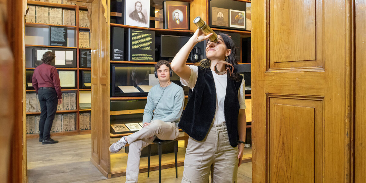 Drei Personen befinden sich in einem Museumsraum mit Holzwänden; eine Person schaut durch ein Fernrohr, eine andere sitzt auf einem Stuhl und eine dritte betrachtet die Ausstellungsstücke.
