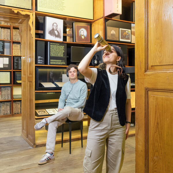 Drei Personen befinden sich in einem Museumsraum mit Holzwänden; eine Person schaut durch ein Fernrohr, eine andere sitzt auf einem Stuhl und eine dritte betrachtet die Ausstellungsstücke.