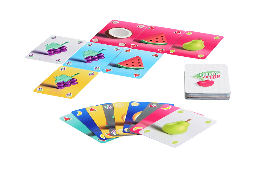 Um ein Kartenspiel mit der Aufschrift „Cherry on Top“ und eine kleine weiße Schale herum liegen mehrere bunte Spielkarten mit Obstmotiven verstreut.