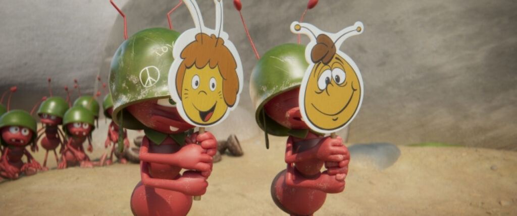 Zwei Comic-Ameisen mit Helmen halten Papiermasken mit lächelnden Gesichtern hoch. Im Hintergrund stehen weitere Ameisen auf einem sandigen Untergrund, hinter ihnen ist eine Felswand zu sehen.