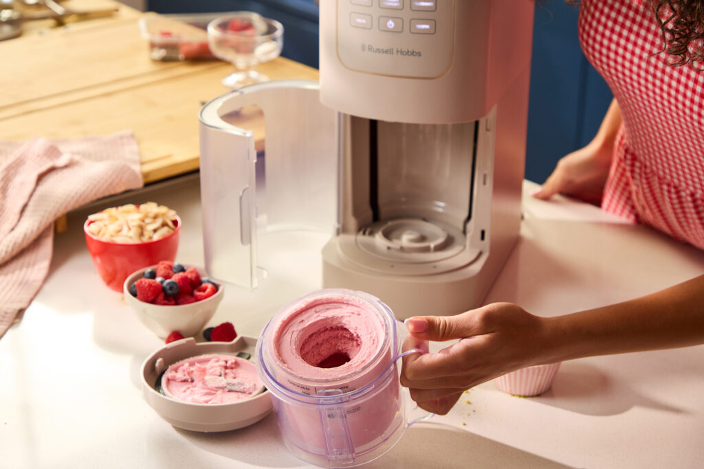 Eine Person hält einen Behälter mit rosa Eis neben einer Eismaschine auf einer Küchenarbeitsplatte, auf der Schalen mit Beeren und Nüssen stehen.