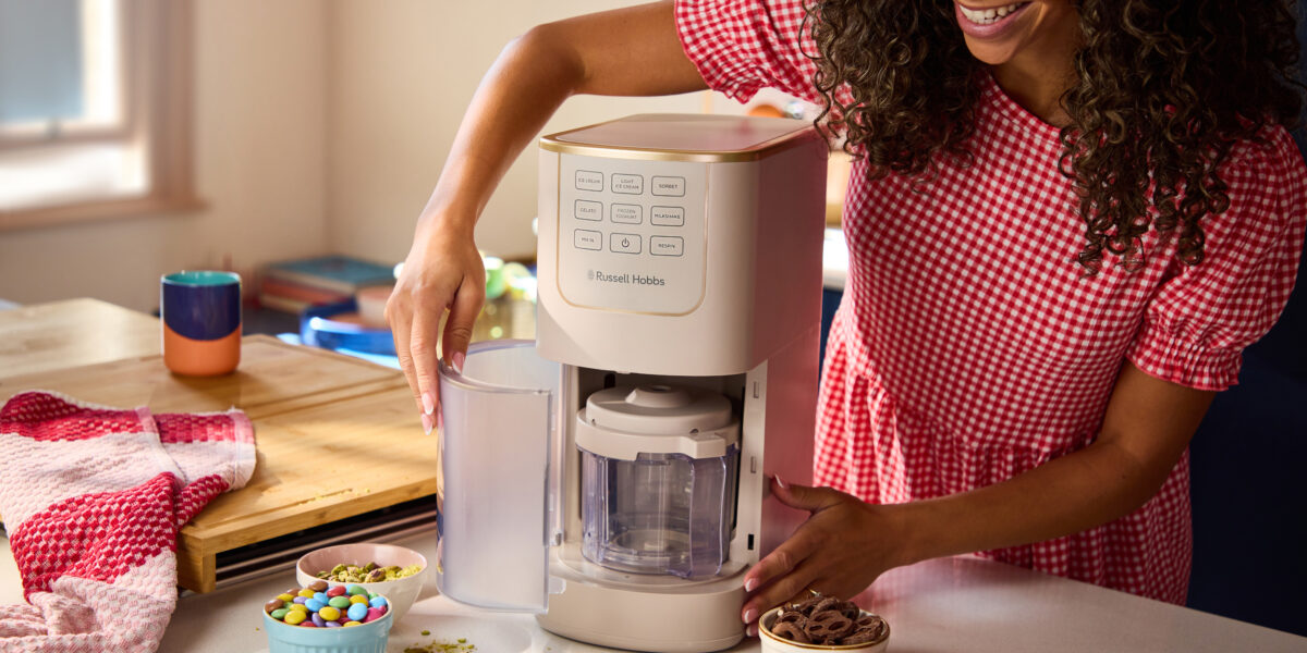 Eine Frau in einem rot karierten Kleid bereitet mit einer Eismaschine von Russell Hobbs ein Dessert zu, umgeben von Schüsseln mit Toppings auf einer Küchenarbeitsplatte.