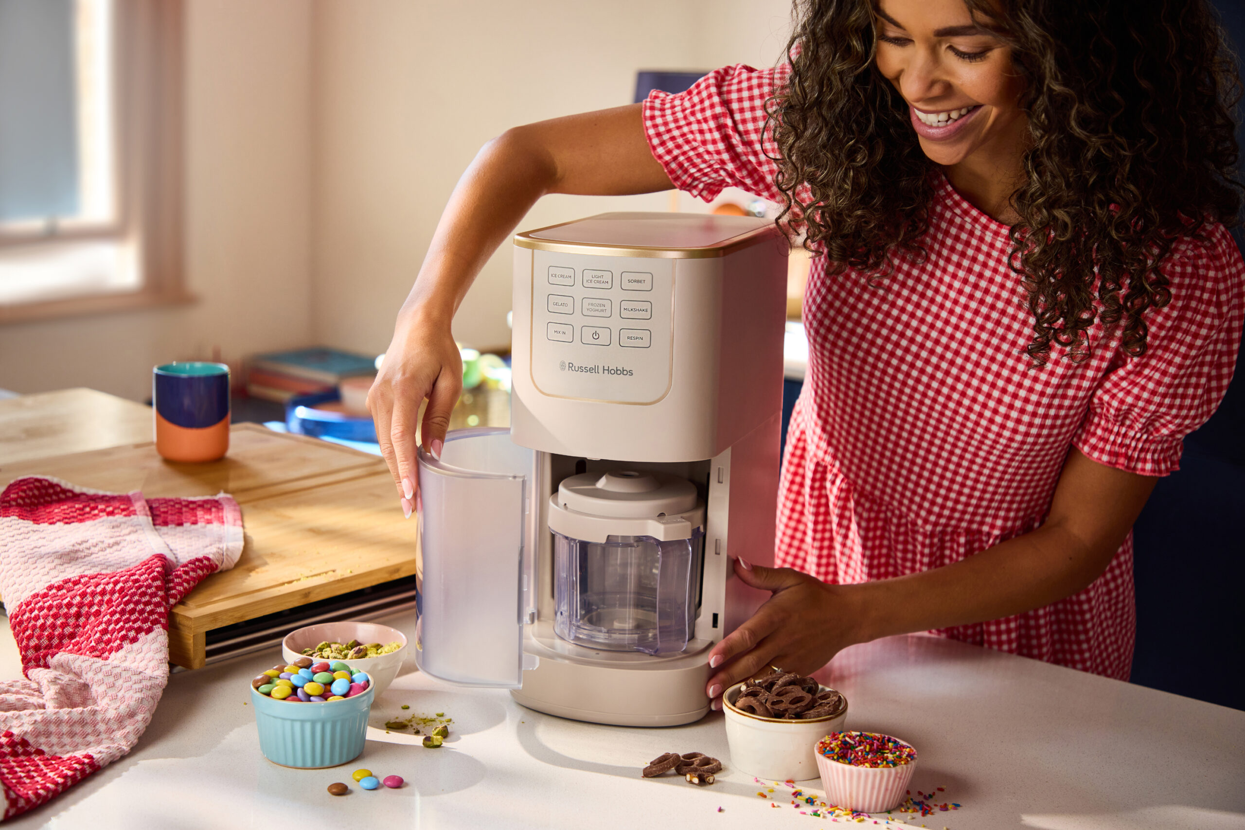 Eine Frau in einem rot karierten Kleid bereitet mit einer Eismaschine von Russell Hobbs ein Dessert zu, umgeben von Schüsseln mit Toppings auf einer Küchenarbeitsplatte.