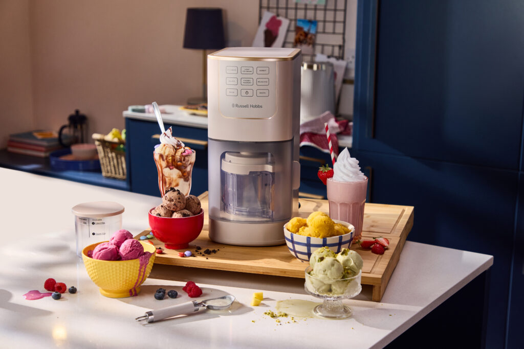 Eine Eismaschine auf der Arbeitsplatte, umgeben von Schalen und Gläsern mit verschiedenen bunten Eissorten und Milchshakes, in einer modernen Küche.