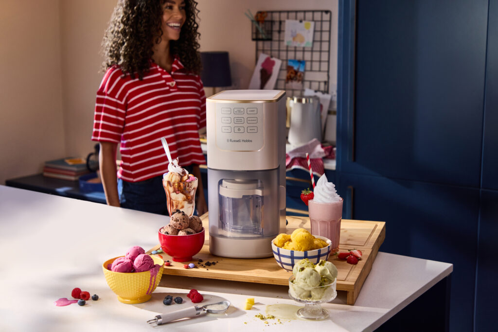 Eine Frau in einem rot gestreiften Shirt steht an einer Theke mit einer Eismaschine, umgeben von Schalen und Bechern mit buntem Eis und verschiedenen Toppings.
