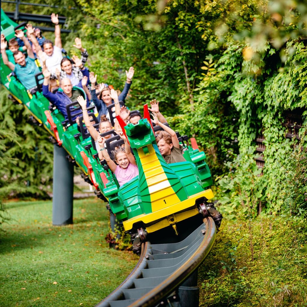 Menschen fahren auf einer Achterbahn mit Drachenmotiv im Freien, umgeben von grünen Bäumen und Gras, wobei viele Fahrgäste bei den Kurven ihre Arme in die Höhe strecken.
