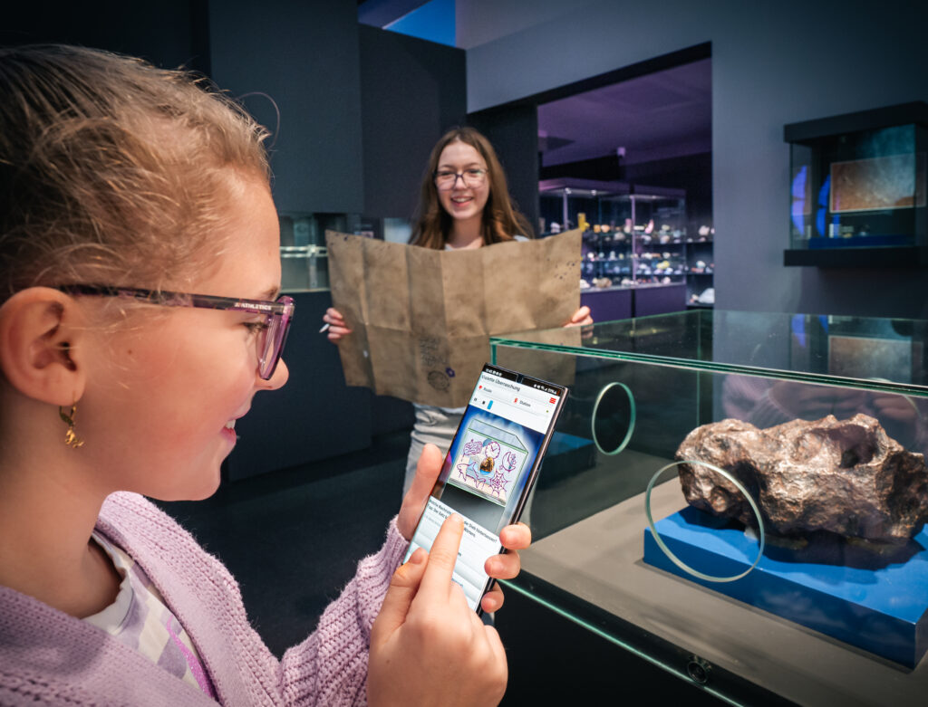 Ein Mädchen mit Brille benutzt ein Smartphone in der Nähe einer Gesteinsausstellung in einem Museum; im Hintergrund hält ein anderes Mädchen eine große Karte in der Hand.
