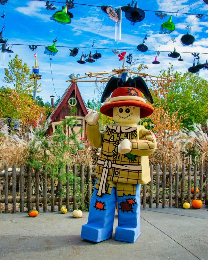 Eine Person in einem LEGO-Kostüm als Vogelscheuche steht im Freien in der Nähe eines Holzzauns, im Hintergrund sind Herbstdekorationen und aufgehängte Hexenhüte zu sehen.