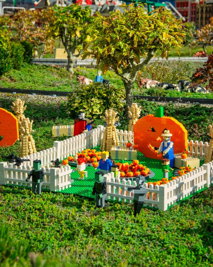 Eine Lego-Szene mit einem Kürbisbeet, einem weißen Zaun, Kürbissen und einer Lego-Figur mit Hut, umgeben von Bäumen und Pflanzen.