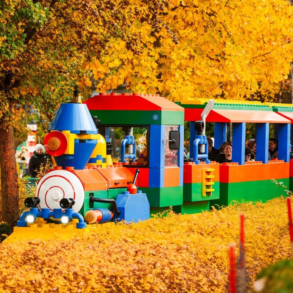 Ein bunter LEGO-Zug fährt durch eine Herbstlandschaft mit goldgelben Blättern und Bäumen und befördert Fahrgäste in offenen Waggons.