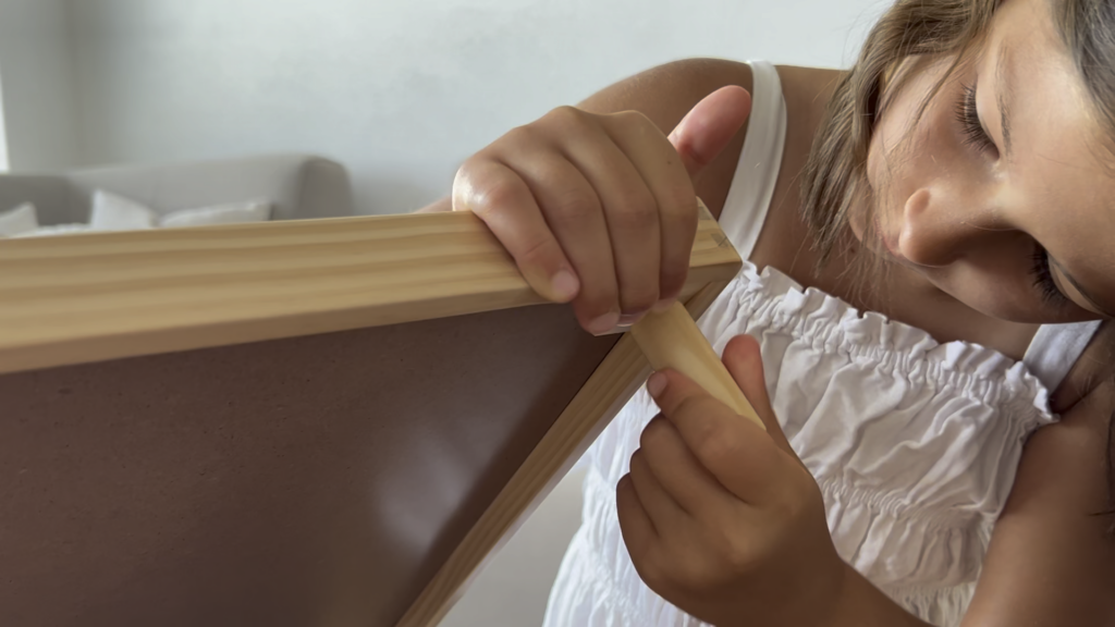 Ein junges Mädchen in einem weißen Kleid baut einen Holzrahmen zusammen und konzentriert sich dabei darauf, einen Stift in die Ecke zu stecken.