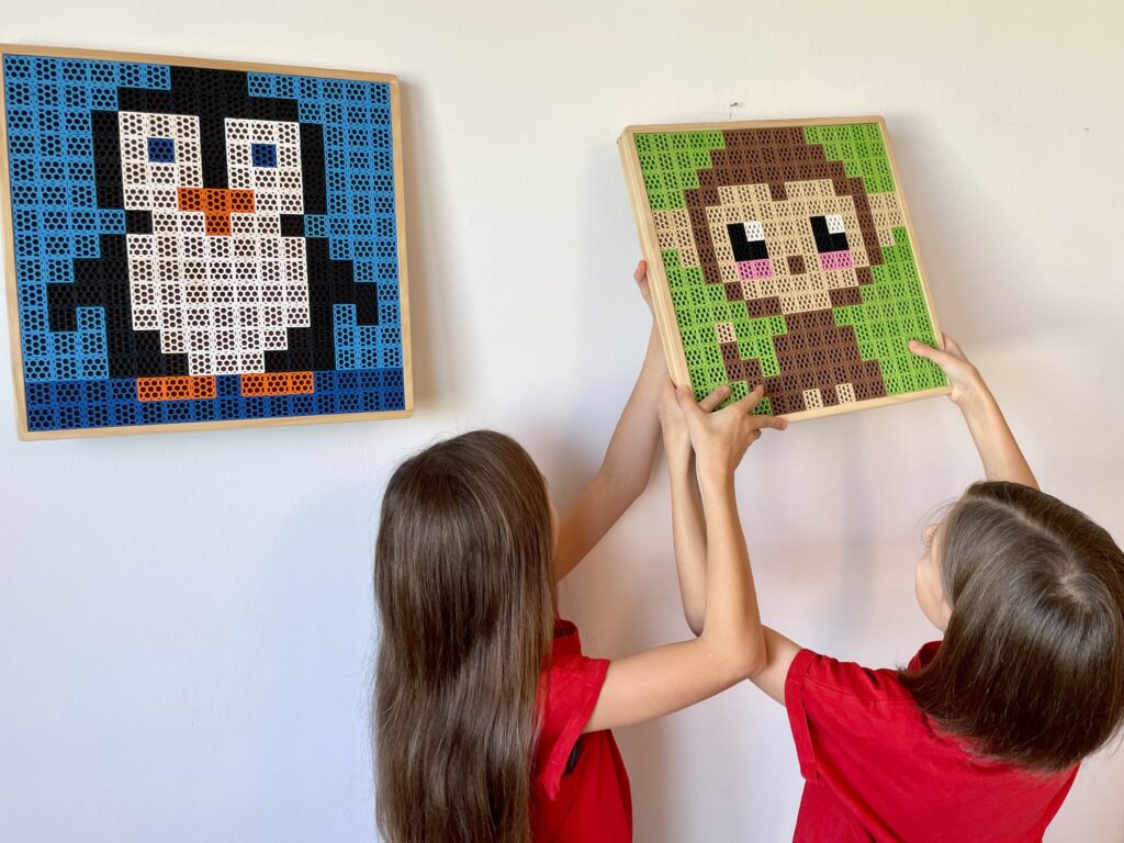 Zwei Kinder in roten T-Shirts hängen ein Pixelkunstbild eines Affen an die Wand, direkt neben ein anderes Pixelkunstbild eines Pinguins.