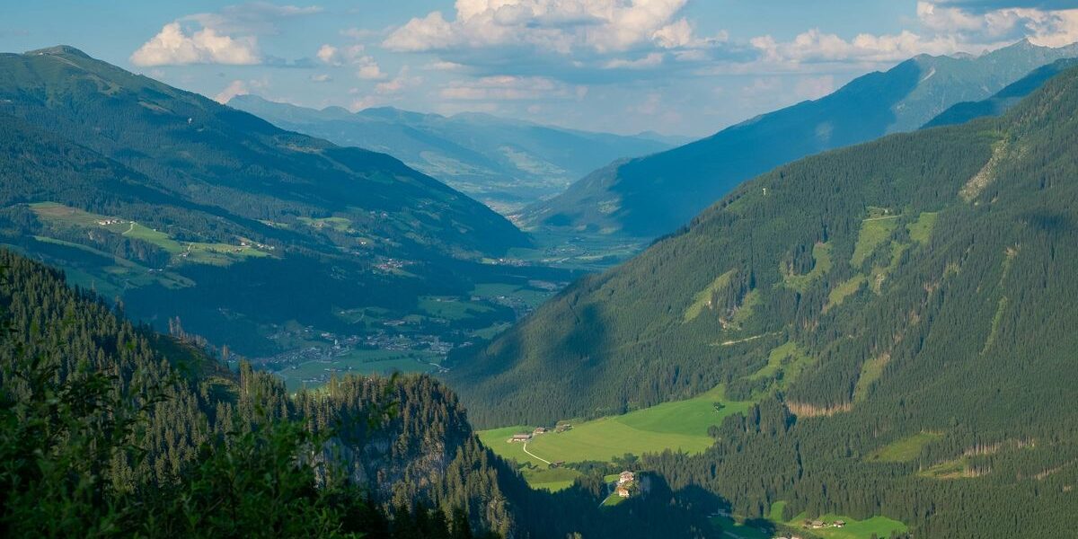 Weitblick auf ein grünes Tal, umgeben von bewaldeten Bergen unter einem teilweise bewölkten Himmel, mit kleinen Gebäuden, die über die Landschaft verstreut sind.