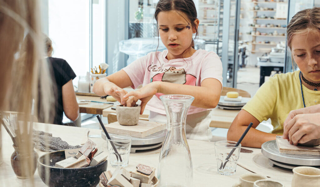 In einem hellen Atelier sitzen Kinder an einem Tisch und töpfern mit Ton und Werkzeugen, umgeben von Schüsseln, Tassen und einem gläsernen Wasserkrug.