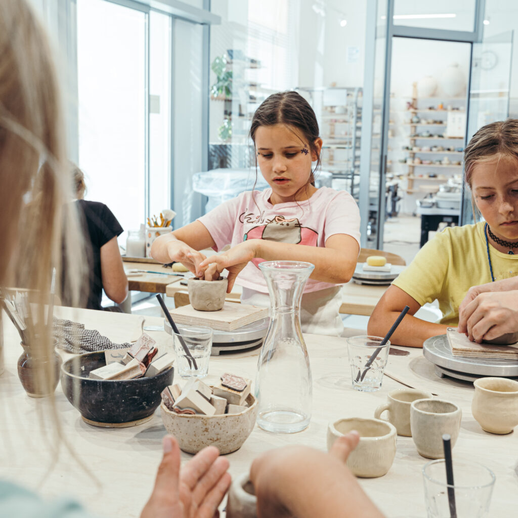 In einem hellen Atelier sitzen Kinder an einem Tisch und töpfern mit Ton und Werkzeugen, umgeben von Schüsseln, Tassen und einem gläsernen Wasserkrug.
