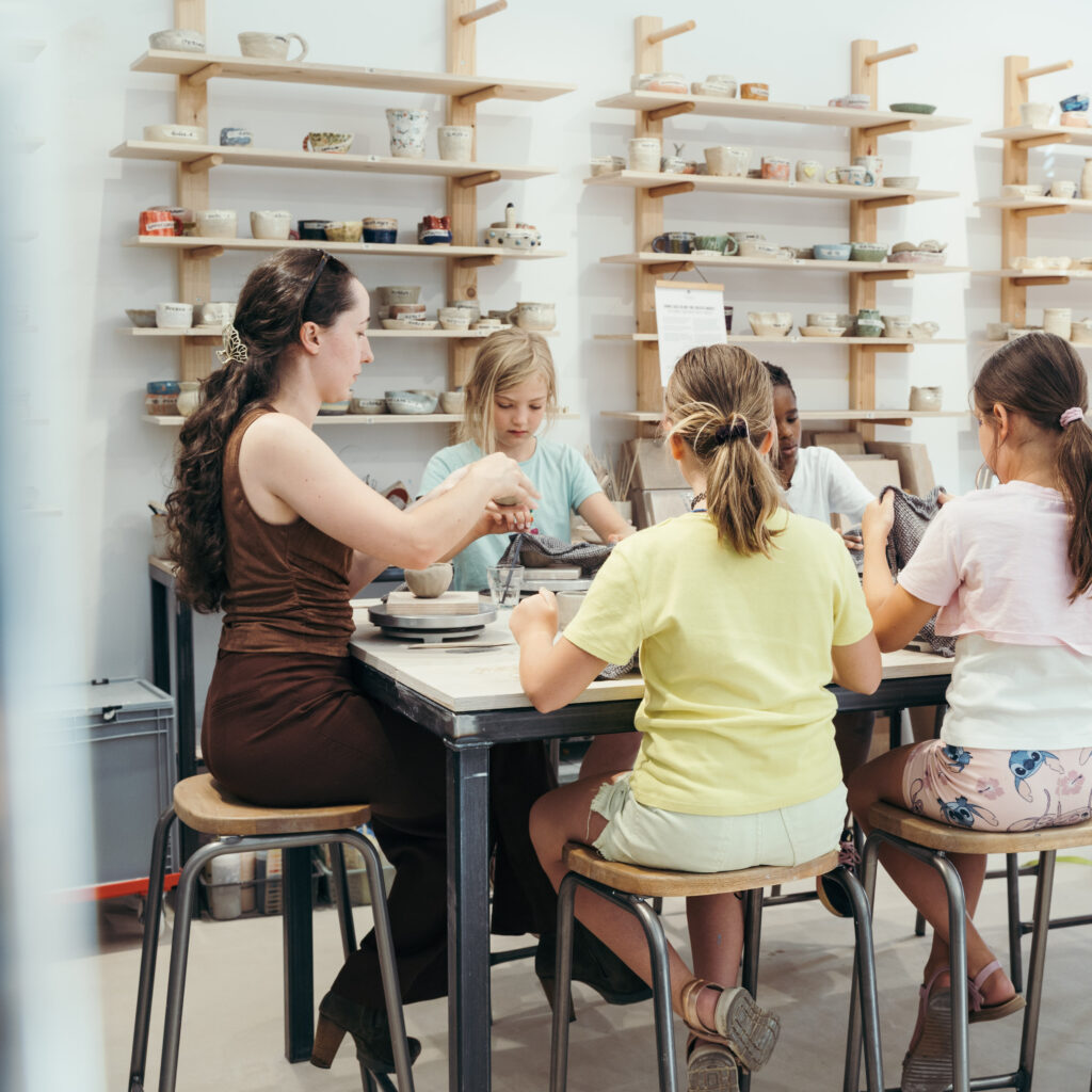 Ein Erwachsener und vier Kinder sitzen in einer Töpferwerkstatt um einen Tisch herum und arbeiten mit Ton, während hinter ihnen an der Wand Regale mit Keramikarbeiten ausgestellt sind.