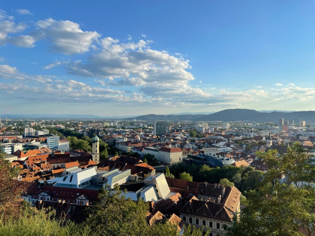 Blick auf eine Stadt mit Gebäuden mit roten Dächern, viel Grün und fernen Bergen unter einem blauen Himmel mit vereinzelten Wolken.