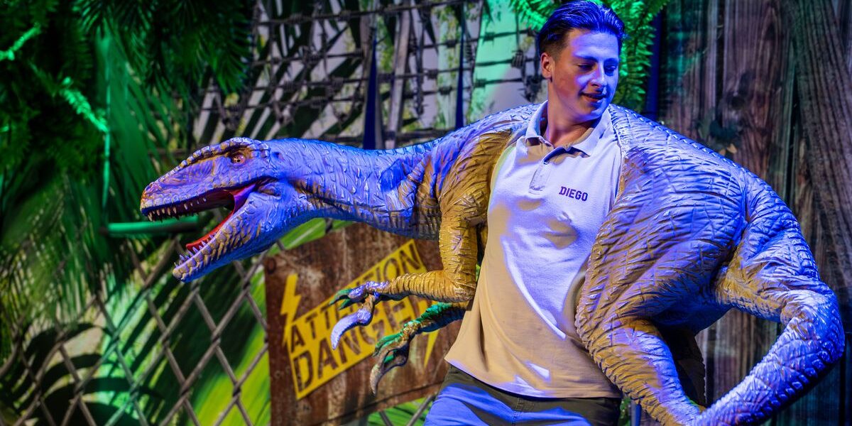 Ein Darsteller namens Diego, der eine blaue Raptor-Puppe in einer Dschungelkulisse mit einem Warnschild im Hintergrund bedient.