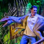 Ein Darsteller namens Diego, der eine blaue Raptor-Puppe in einer Dschungelkulisse mit einem Warnschild im Hintergrund bedient.