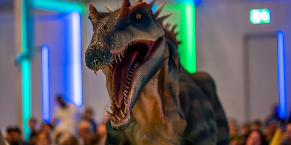 Eine realistisch wirkende Dinosaurier-Animatronik mit geöffnetem Maul steht vor dem Publikum in einem Raum, der von blauem und grünem Licht beleuchtet wird.