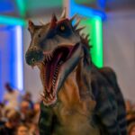 Eine realistisch wirkende Dinosaurier-Animatronik mit geöffnetem Maul steht vor dem Publikum in einem Raum, der von blauem und grünem Licht beleuchtet wird.