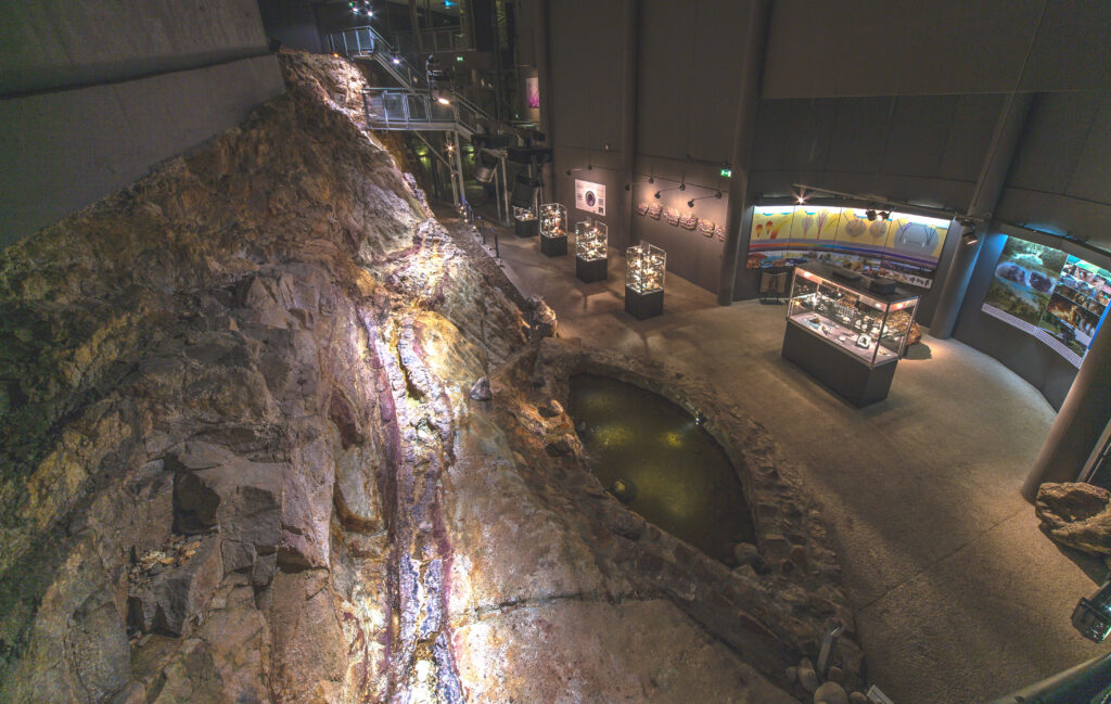 Eine Museumsausstellung umfasst eine große freiliegende Felsformation, einen kleinen Innen-Teich, Vitrinen mit Exponaten sowie Informationstafeln, die in sanftes Licht getaucht sind.